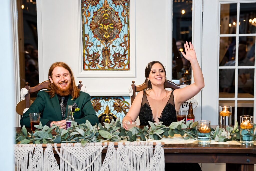 Parker and Olivia seated at their reception table while Olivia raises her hand