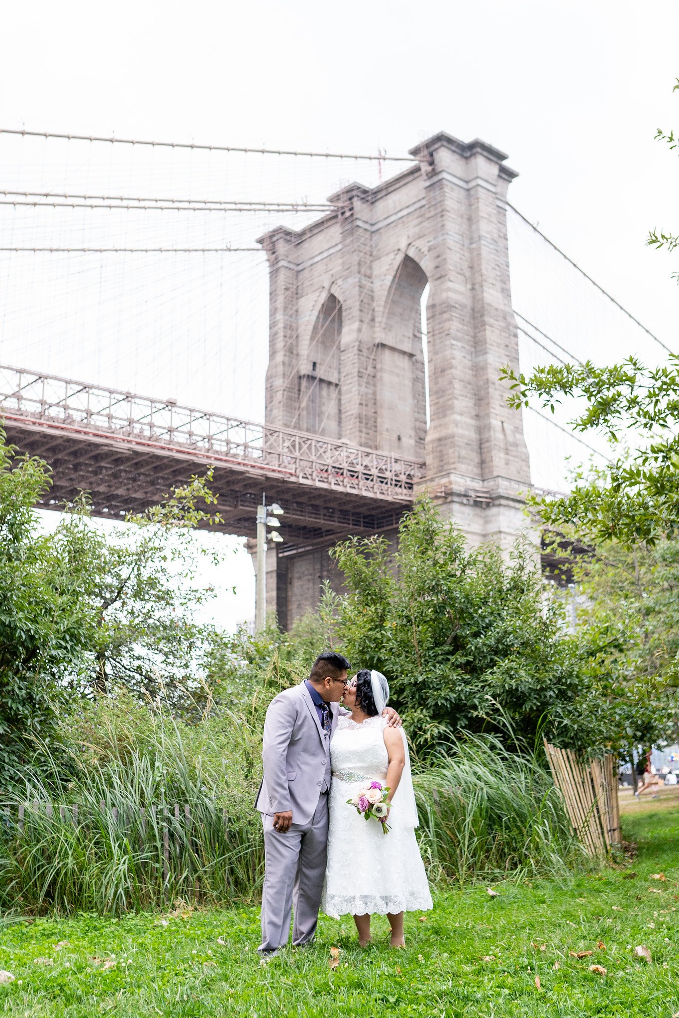 The couple stand among greenery beneath a large suspension bridge.
