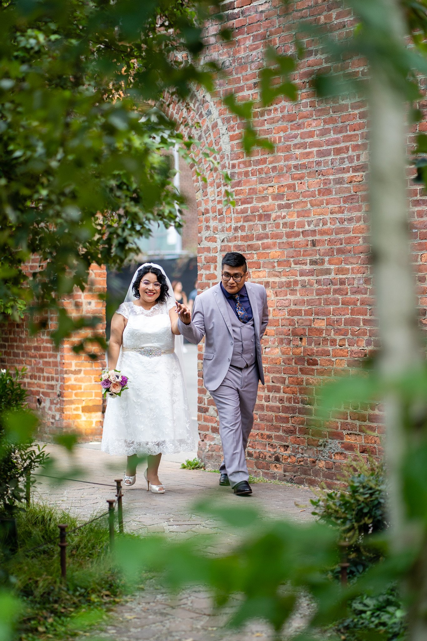 The newlyweds walk together, framed by leaves in the foreground.
