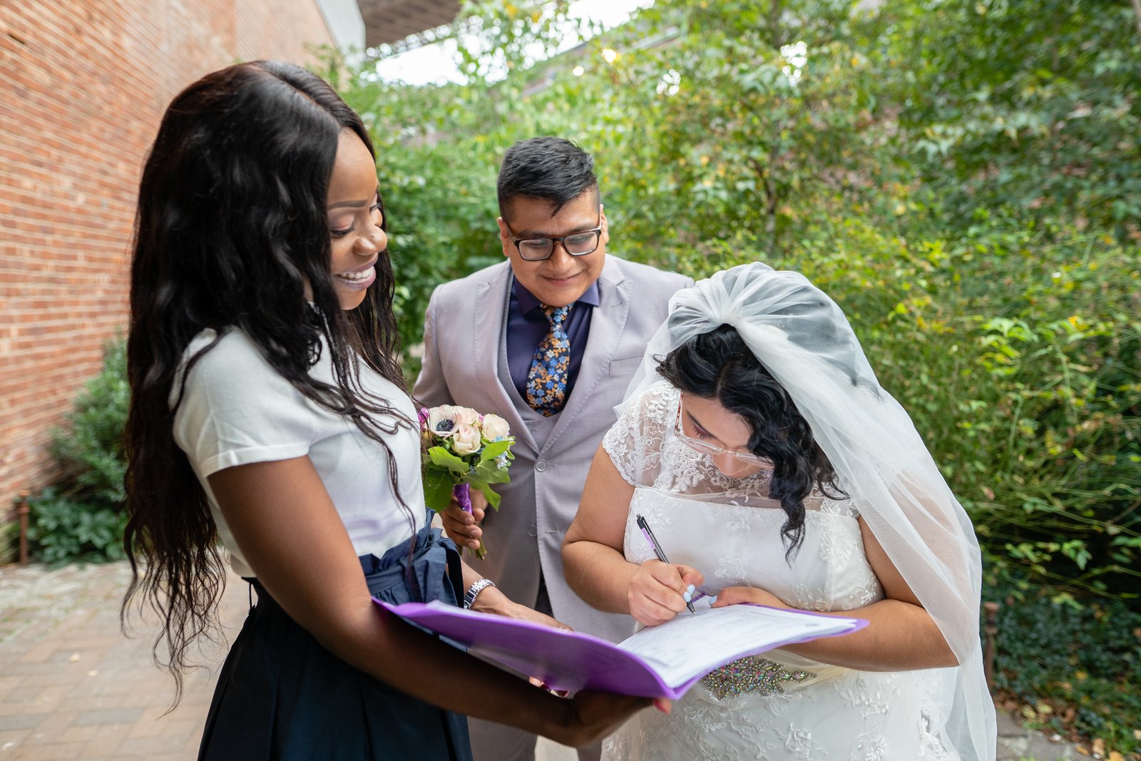 Brenda signs the marriage paperwork as David and the officiant look on.