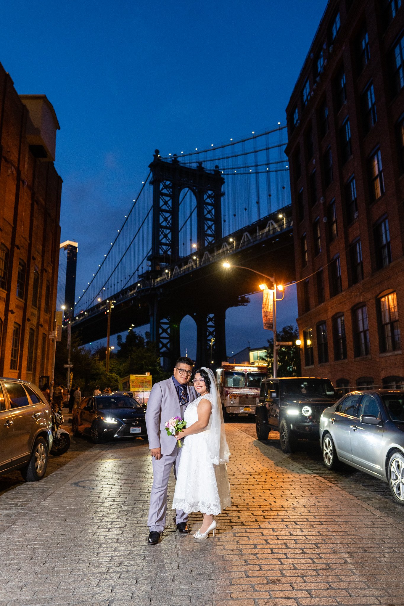 The newlyweds stand on a city street beneath a bridge at night.