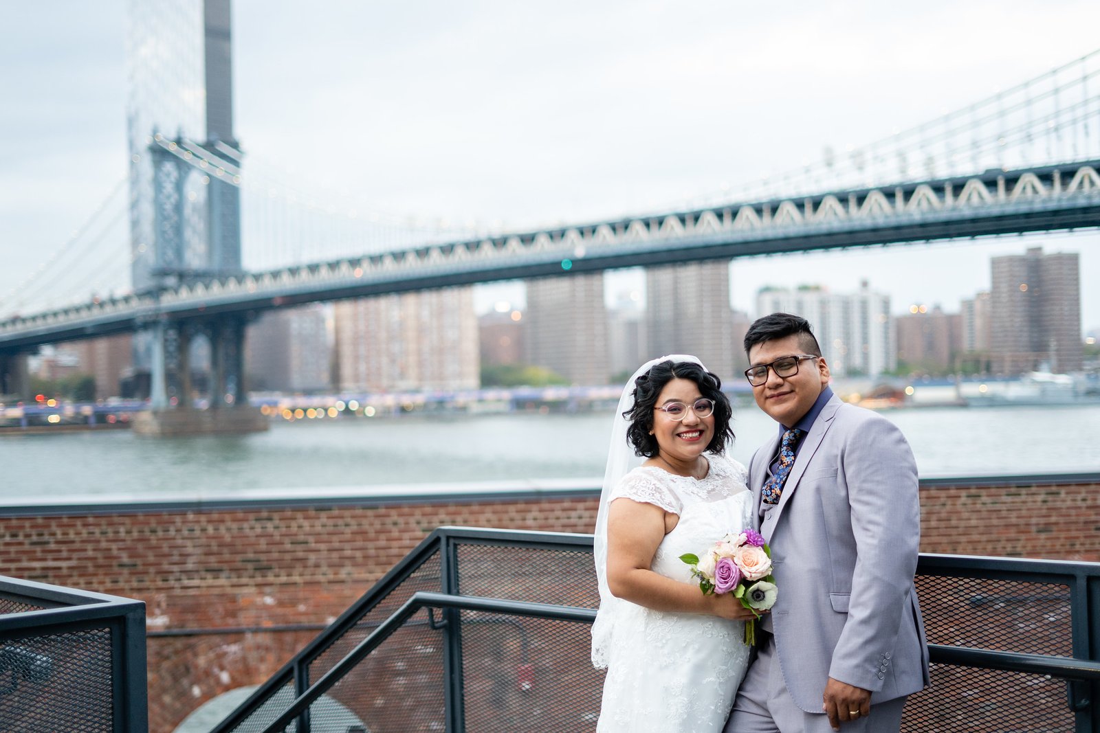 The newlyweds stand beside the river with a bridge behind them.