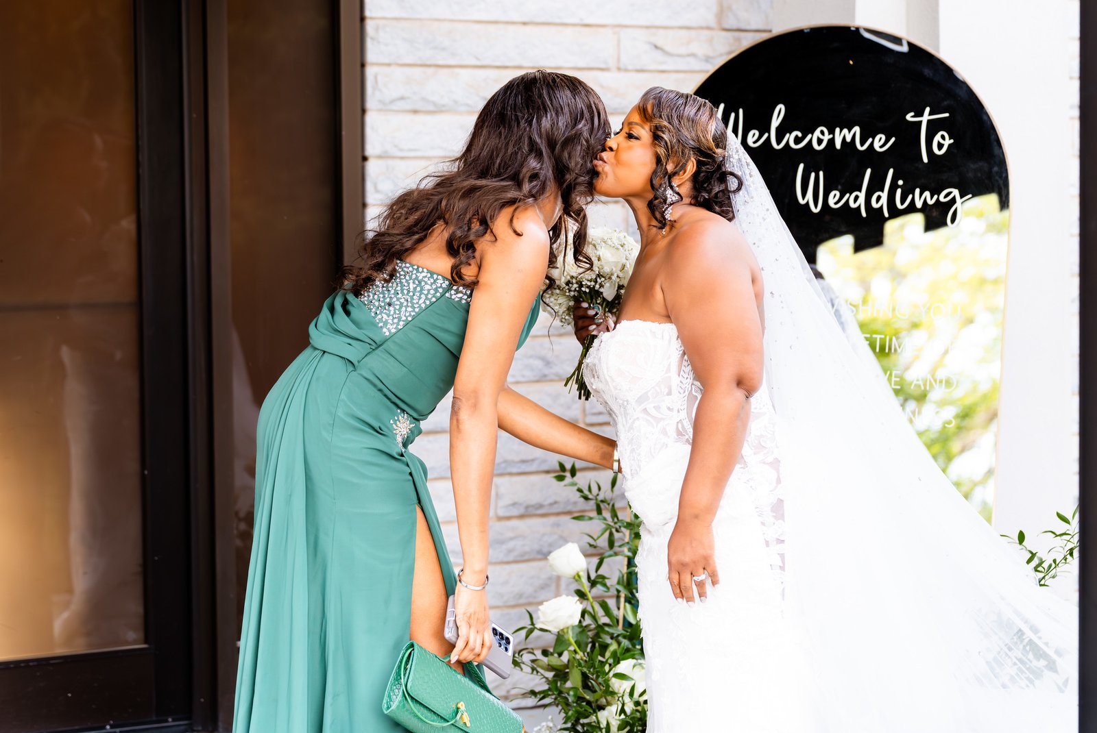 The bride kisses a guest in a green dress beside a wedding welcome sign.