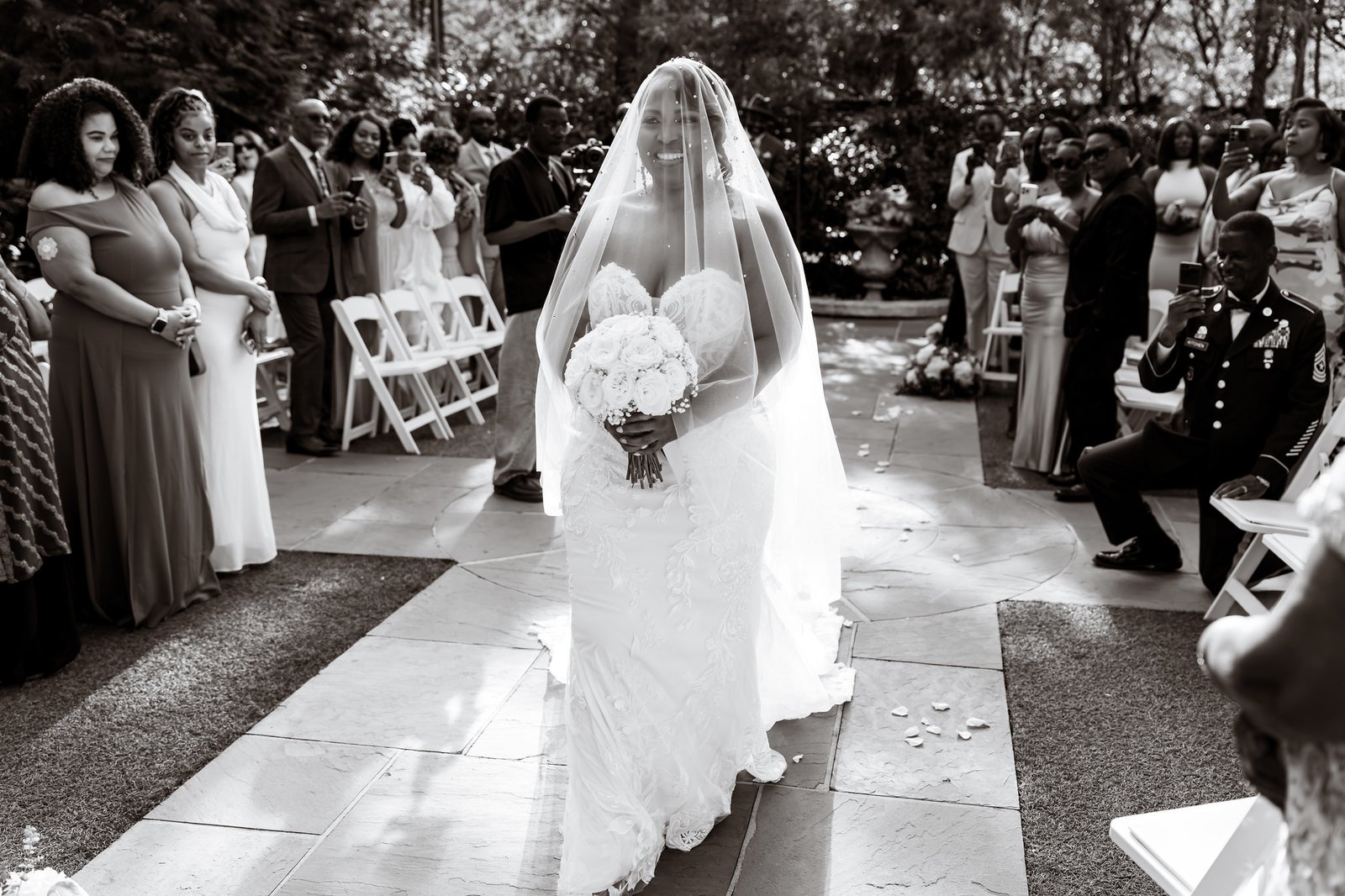The bride walking down the aisle beneath her veil as guests watch.