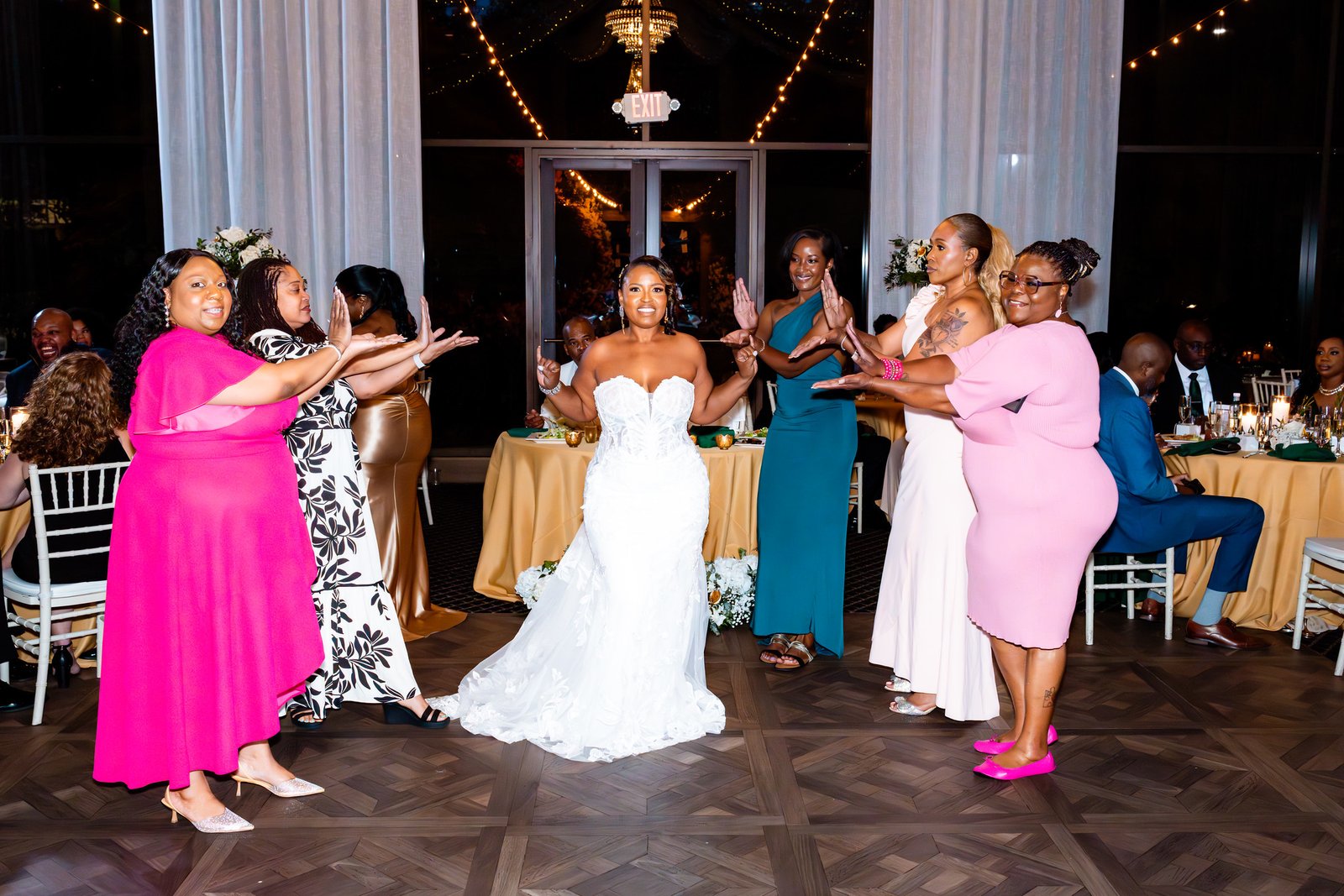 Guests in colorful dresses gesture toward the smiling bride on the reception floor.