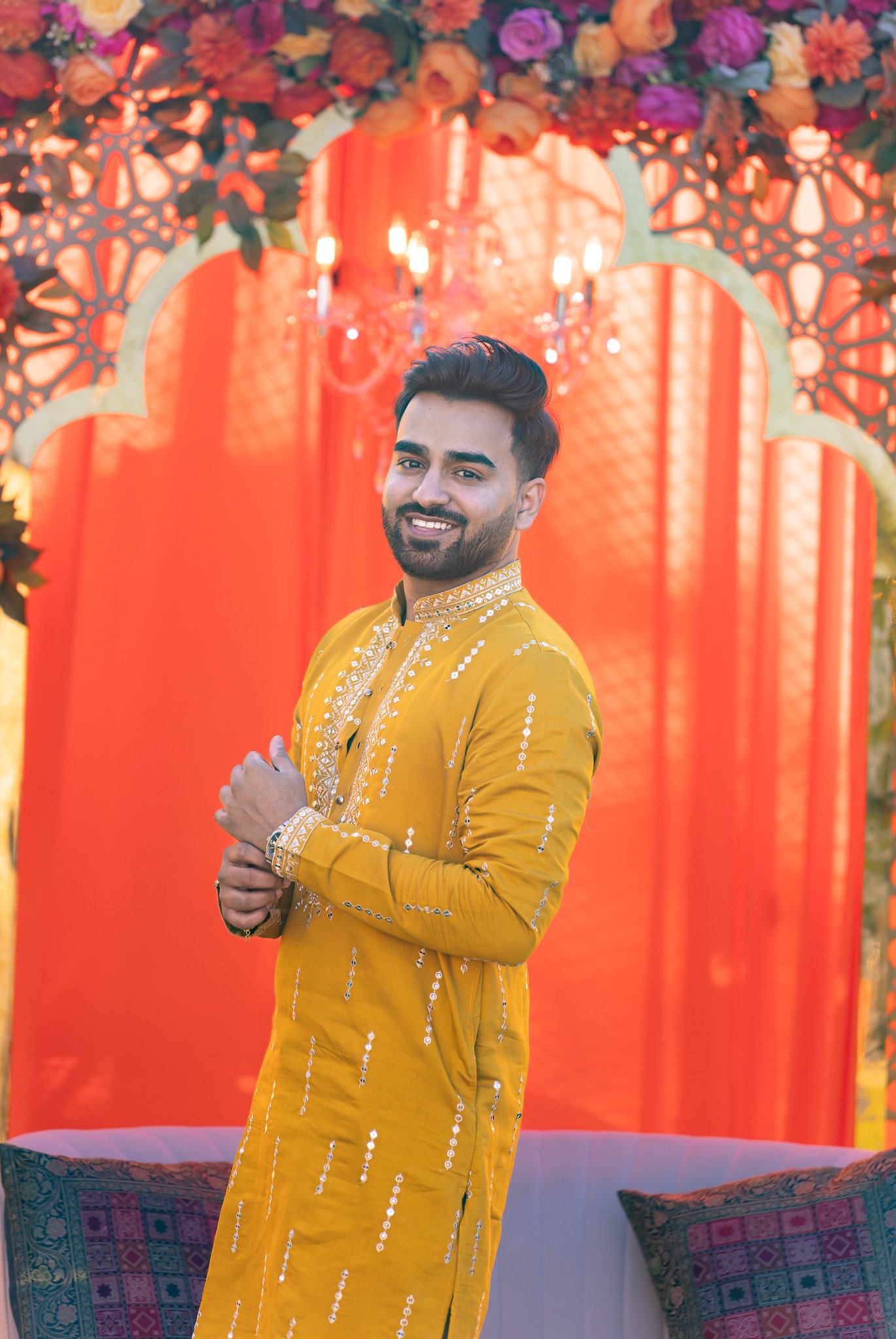 Groom smiles in an embroidered yellow outfit against an orange backdrop.