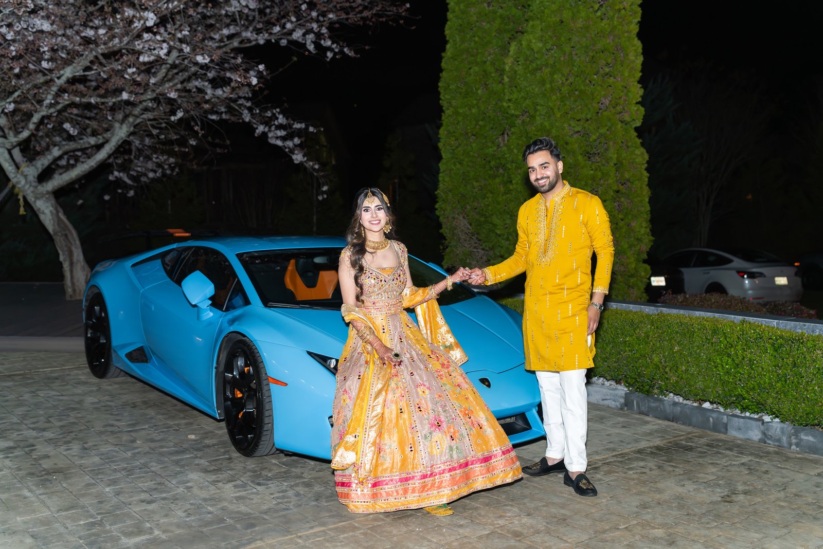 A couple holding hands beside a blue sports car at night.