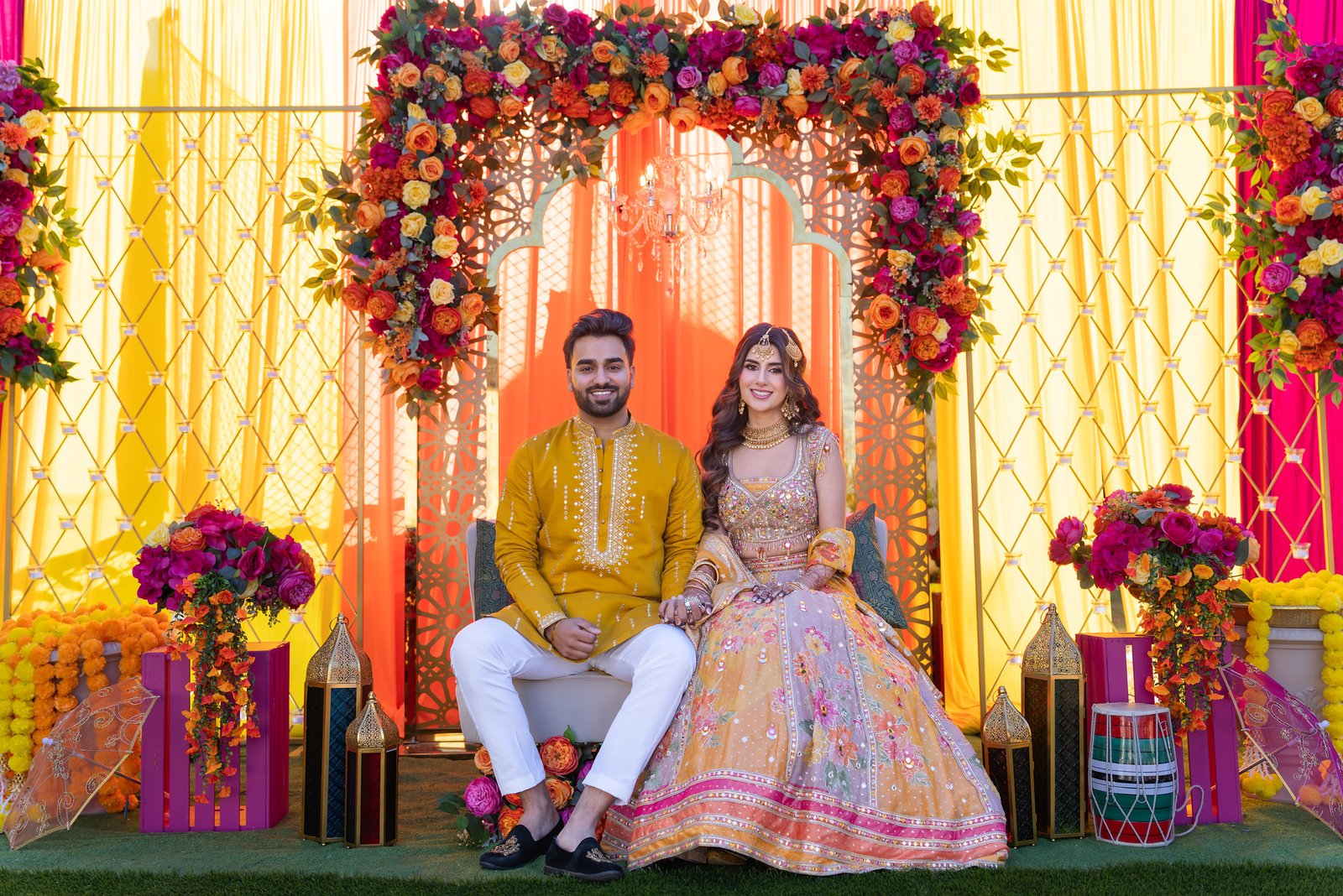 Couple seated beneath an orange and yellow floral backdrop, smiling and holding hands.