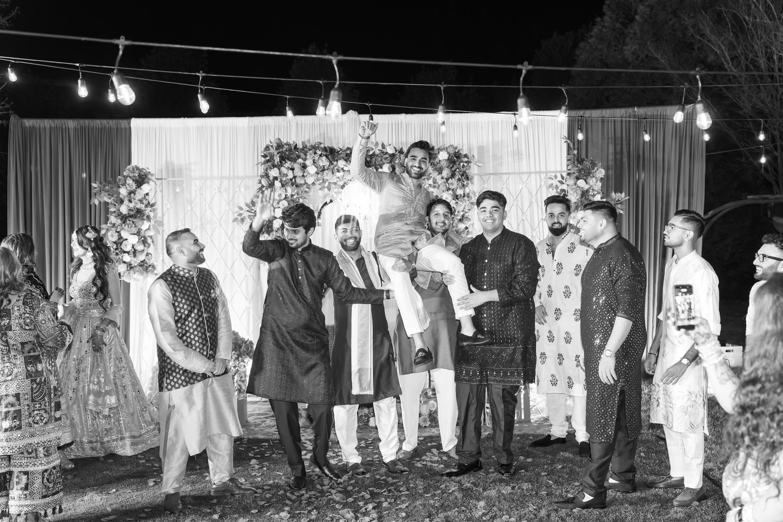 Guests lift a smiling man into the air beneath string lights in a black-and-white photograph.