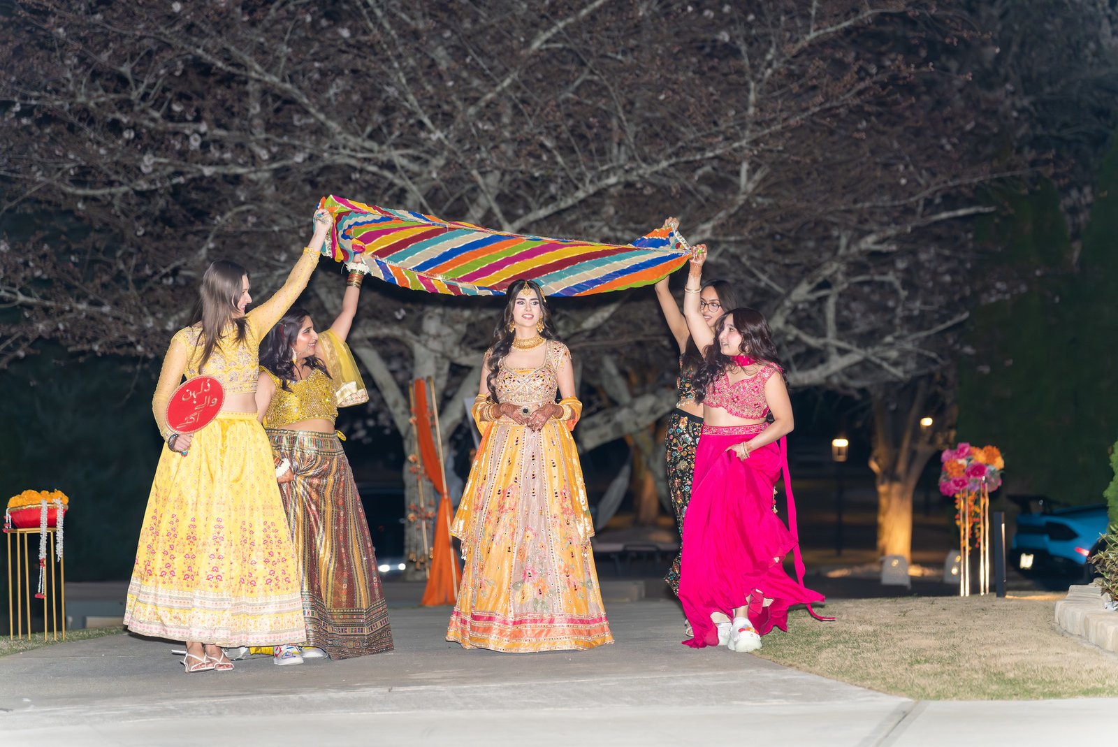 Bride walks with guests holding a colorful fabric canopy above her at night.