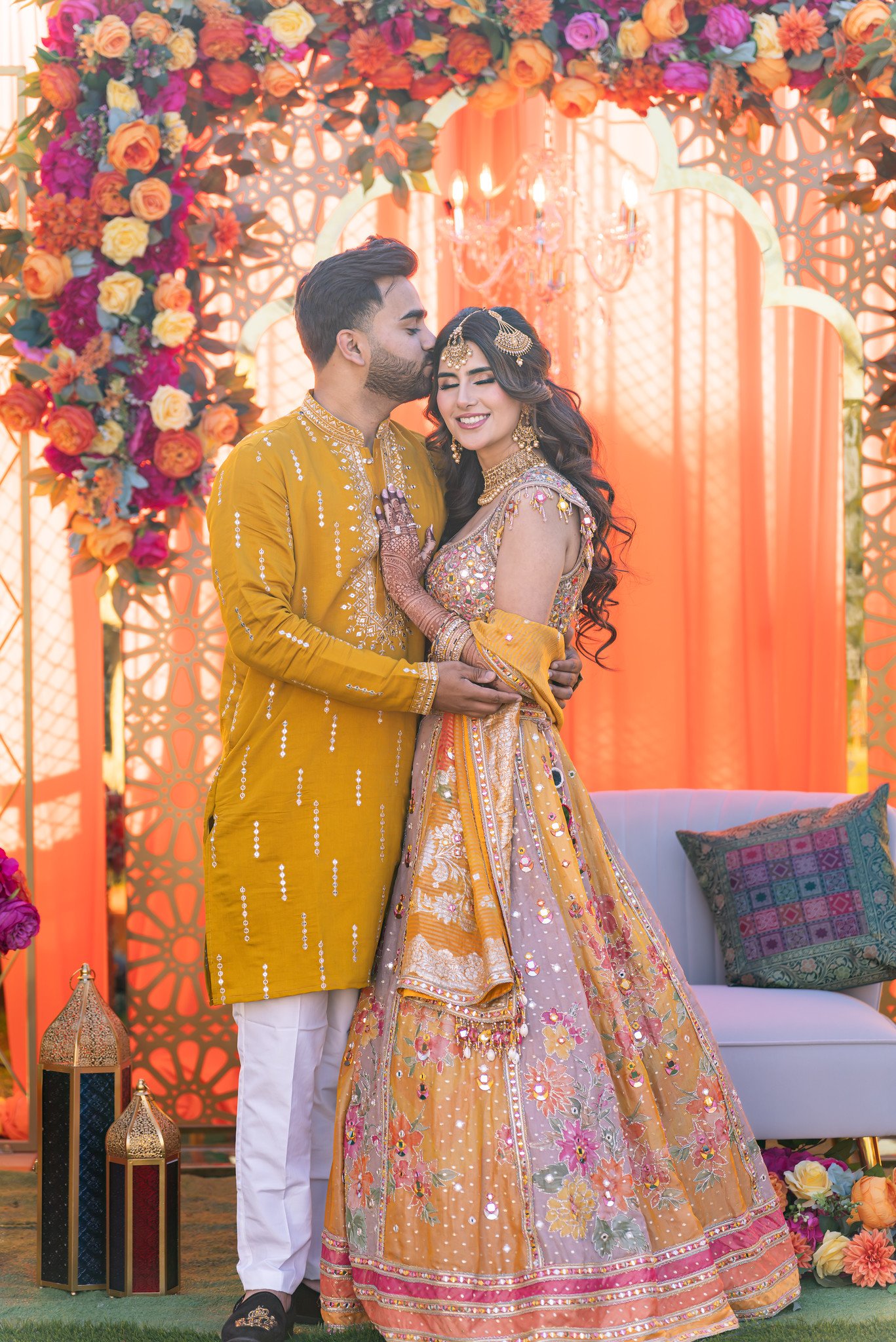 Groom kisses the bride's forehead as she smiles against a bright floral backdrop.