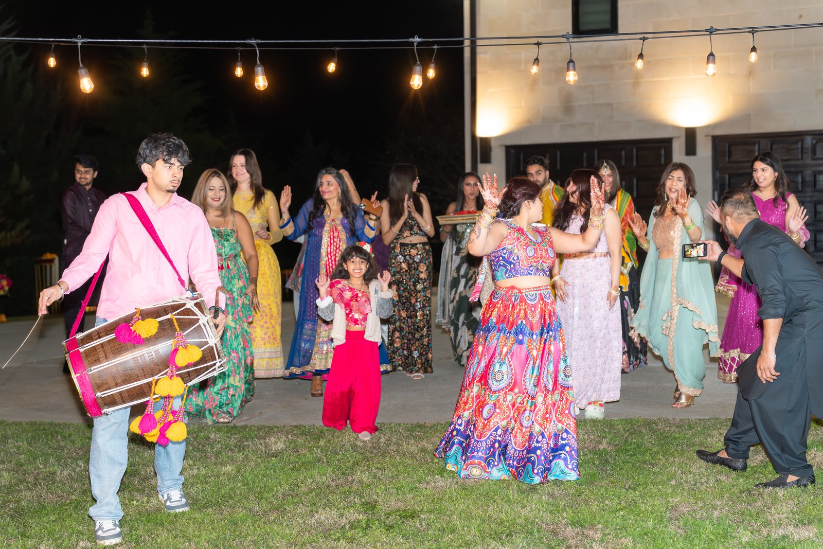A drummer leads dancing guests outdoors beneath string lights.
