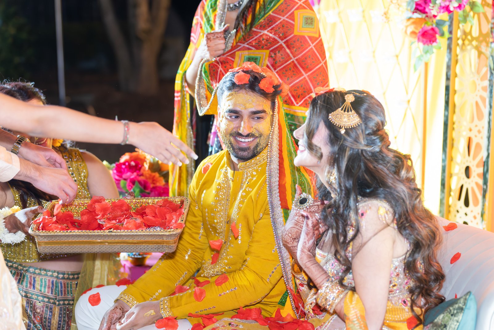 A smiling couple seated together with red petals and guests’ hands in the foreground.