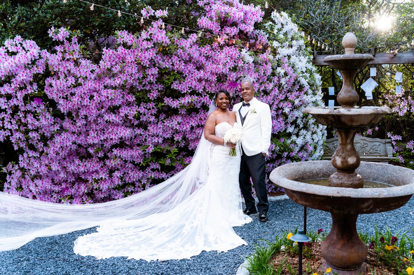 The newlyweds stand beside a wall of pink flowers and a stone fountain, with the bride’s train spread across the path.